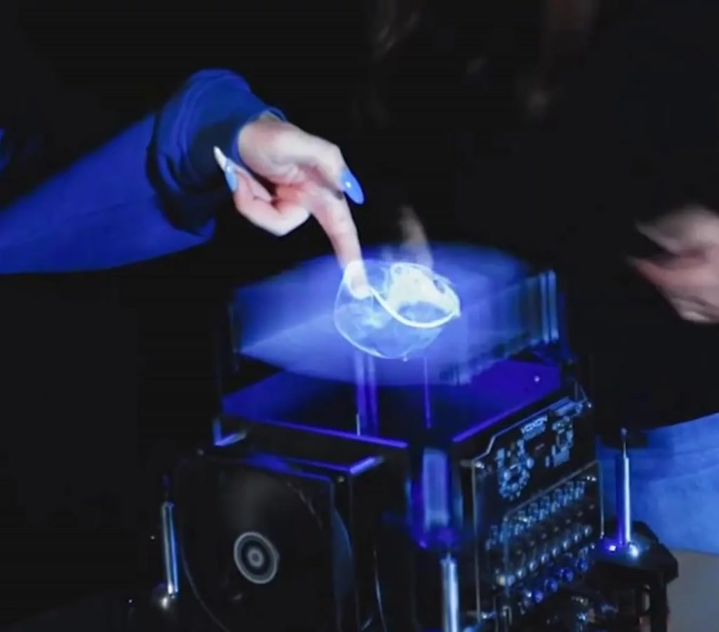 Person inserting a right hand into a vertical glowing 3D hologram sphere with a wrist-mounted haptic device and visible projection hardware