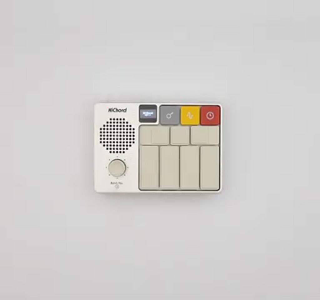 Vertical photo of the Pocket Audio HiChord handheld controller showing seven backlit buttons and a central joystick held above mixing desk faders